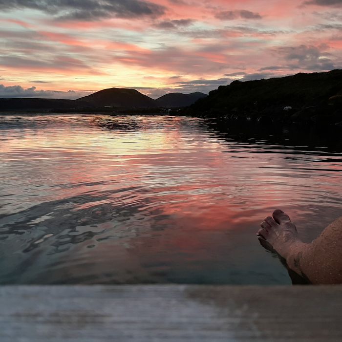 Ein Sonnenaufgang hinter einem Berg, davor der große See mit nackten Füßen, die ins Bild ragen