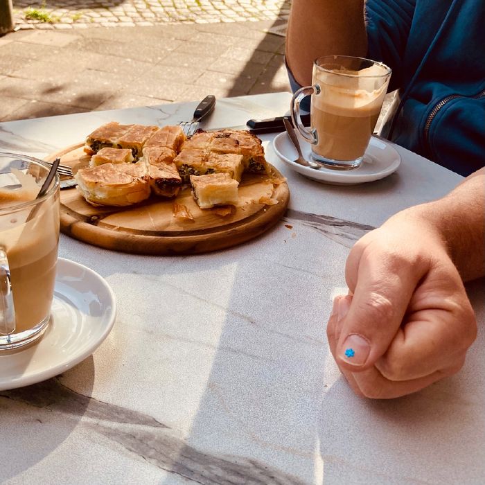 Blick auf einen Tisch mit 2 Gläsern mit Milchkaffee und Börek. Ein Daumen wird in Richtung Kanera gehalten, auf dem Fingernagel ein Blümchen-Sticker