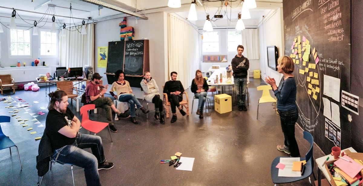 Gruppenpanorama in der tollwerkstatt. Kai, Bertram, Judith, Annika, Jeff, Nina und Klaus sitzen / stehen im Kreis vor der Tafel. Natalie steht und spricht zur Runde, hinter ihr Post-Its und Texte an der Tafel. Fotografiert von Joschi.