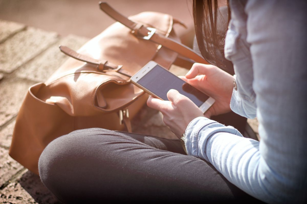 Nahaufnahme der Hände einer Frau, die auf dem Boden sitzend ein Android-Smartphone in der Sonne nutzt
