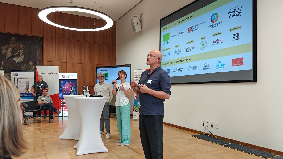 Johannes Bisping, Dorothee Brommer und Tom Ritschel bei der Eröffnung der SustaiNable Conference 2025 in den Räumen der IHK Nürnberg. Doro Brommer spricht in ein Mikrofon, im Hintergrund werden die Logos der Sponsoren projiziert.