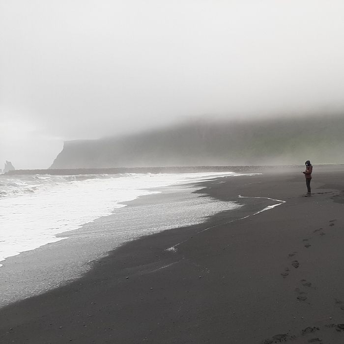 Nina steht rechts am schwarzen Strand und blickt aufs Meer