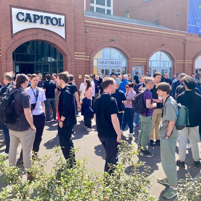 Vor einem roten Backsteingebäude mit dem Schild „Capitol“ stehen viele Menschen in der Sonne und unterhalten sich