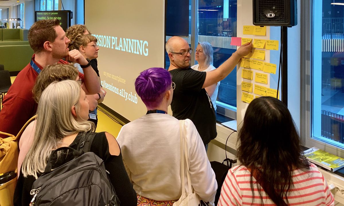 Umgeben von ein paar Personen steht ein Mann vor einer Wand mit Post-its. Er ist dabei, die Position eines der gelben Zettel zu ändern, während er sich gleichezitig der Gruppe zuwendet.