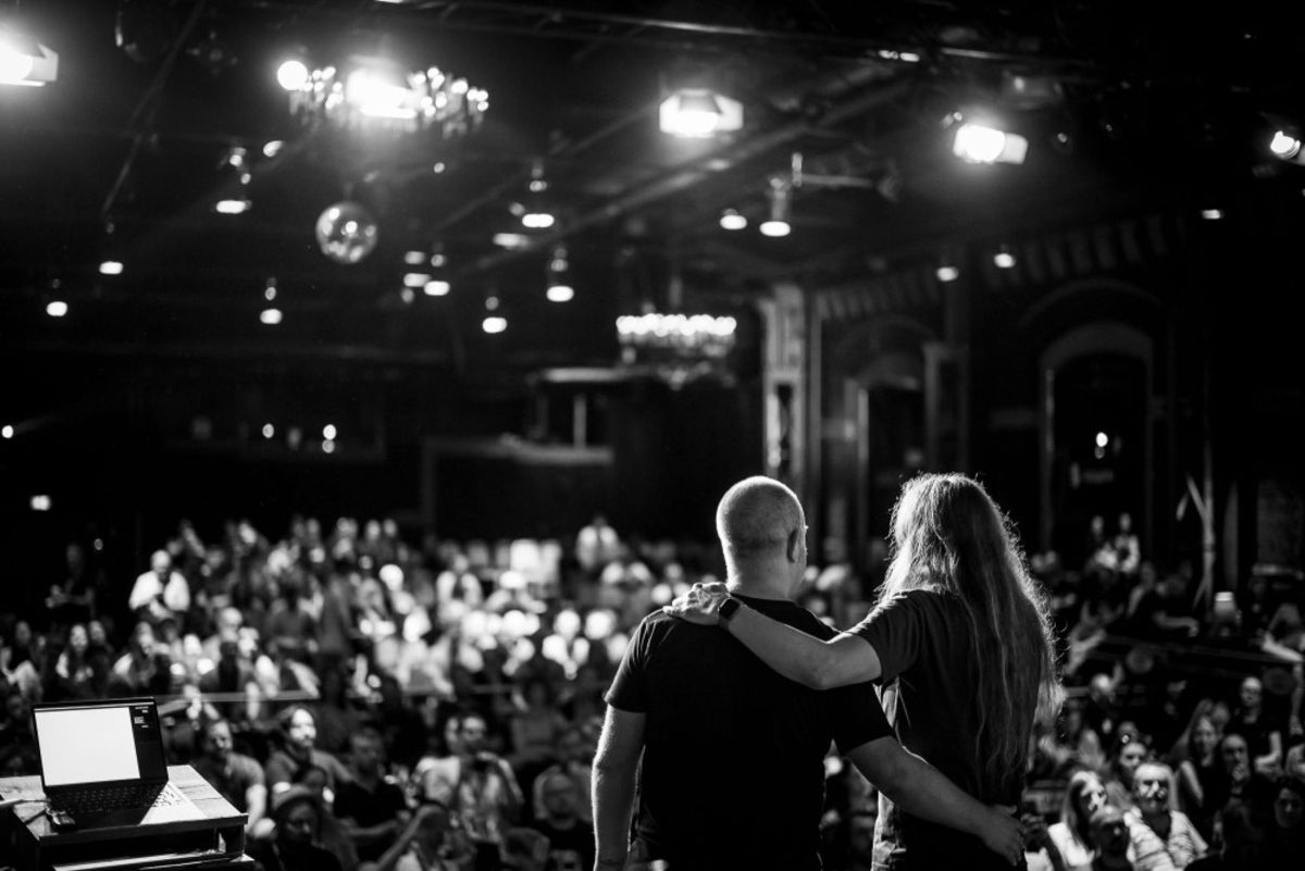 Schwarz-Weiß-Foto von der Bühne über den Publikumsraum. Joschi und Marc stehen mit dem Rücken zur Kamera und haben die Arme um die Schultern gelegt.