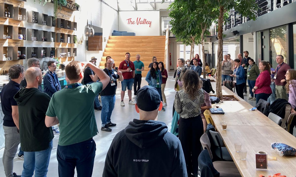 In einem großen Raum steht ein Mann mit schwarzem T-Shirt und spricht zu einer großen Gruppe von Personen, die ihn umgeben.