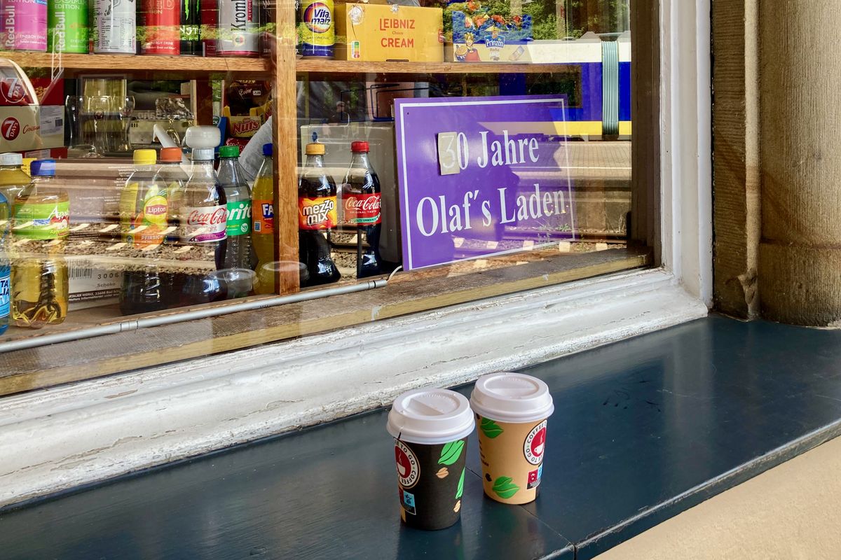 Auf einem Fenstersims stehen zwei Kaffee-to-go. Durch das Fenster sieht in das Innere, im Regal stehen Getränkeflaschen, andere typische Kioskprodukte und ein Schild "30 Jahre Olafs Laden"
