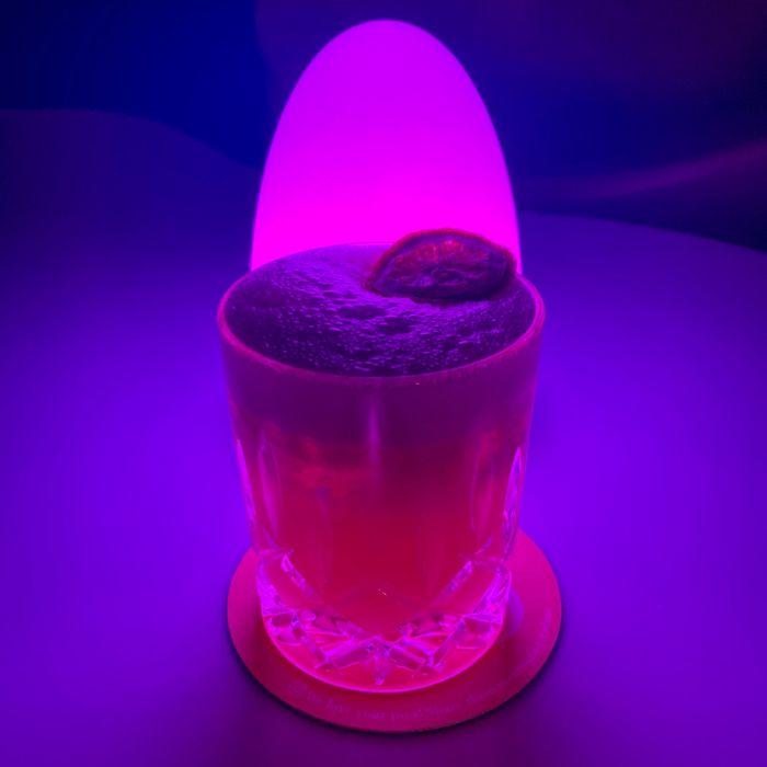 Ein Cocktail mit Eiweißschaum und einer getrockneten Orange steht vor einer rosa schimmernden Lampe.