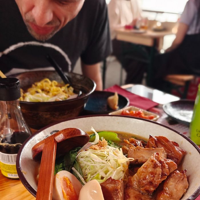 Im Vordergrund eine Schale Teriyaki Miso Ramen. Im Hintergrund unscharf beugt sich David über seine Schale.