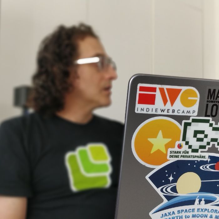 Tantek Çelik hinter seinem aufgeklappten Laptop. Er trägt ein Microformats-T-Shirt. Auf seinem Laptop kleben viele Aufkleber, darunter ein IndieWebCamp-Sticker