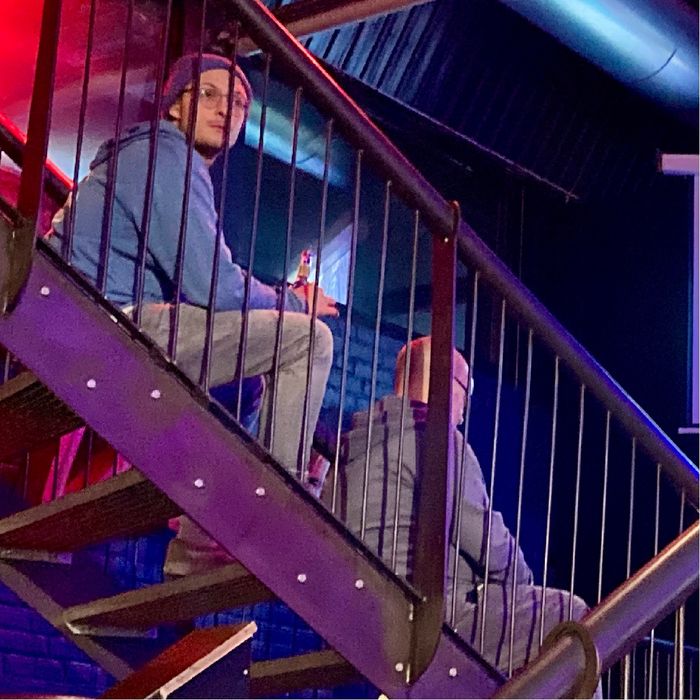 Zwei Männer sitzen hintereinander auf einer Stahltreppe, der hintere schaut in Richtung Kamera. Beide sind von der Seite zu sehen