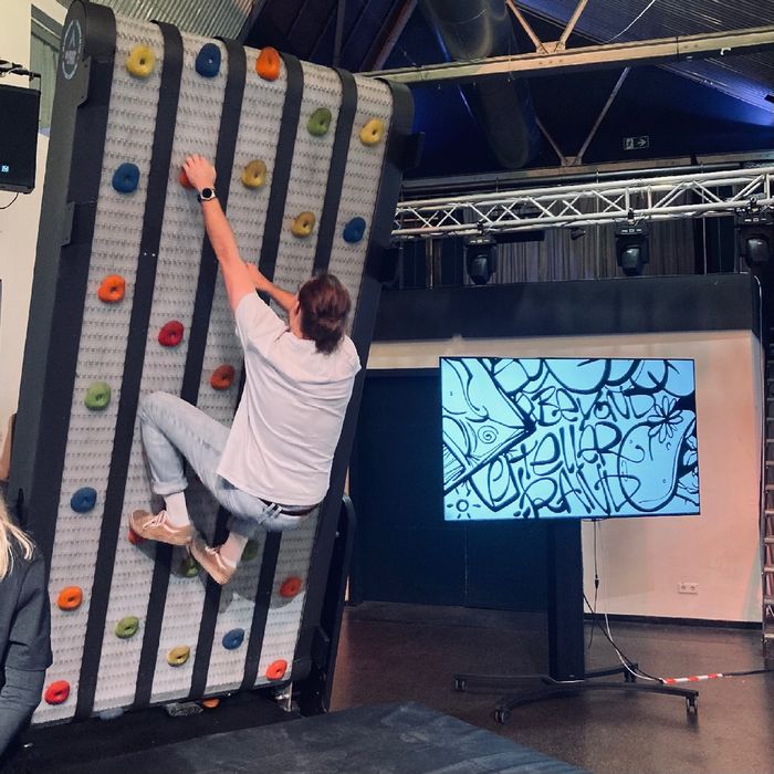 Jakob hängt an einer Kletterwand, die um einige Grad nach vorne gekippt ist.