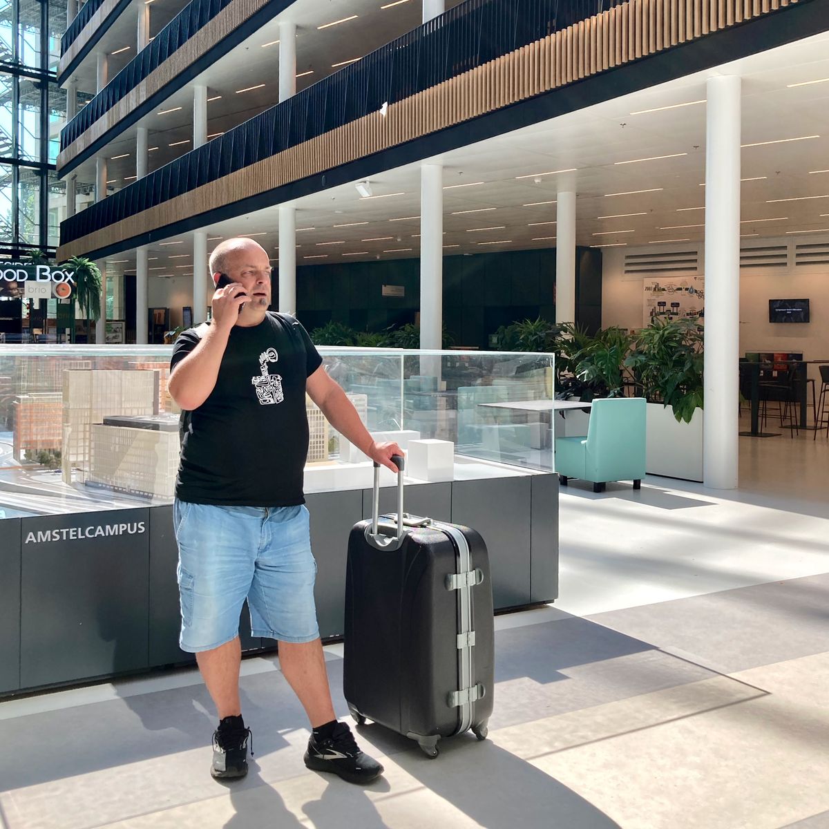 Ein Mann in kurzen Hosen und schwarzem T-Shirt steht in einer modern anmutenden Lobby und telefoniert. Er hat einen großen Koffer neben sich stehen.