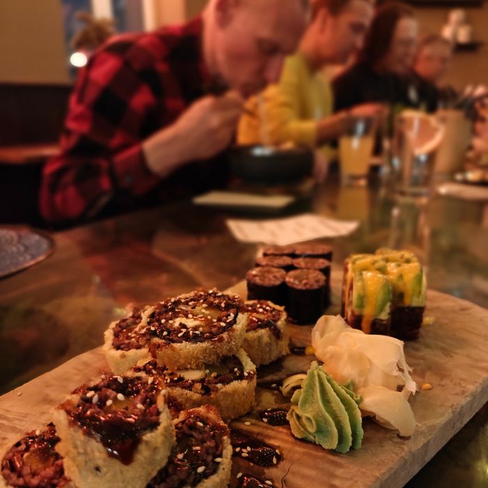 Auswahl von lecker angerichtetem veganen Sushi auf einem Essbrett. Im Hintergrund unscharf Guido und Marc beim Essen