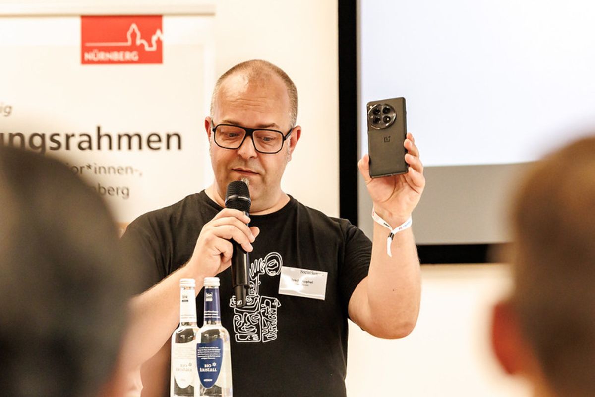Joschi Kuphal beim Vortrag seiner These. In der linken Hand hält er ein Mikrofon, mit der rechten Hand hält er sein Smartphone in die Luft. Sein Blick ist nach unten gerichtet.