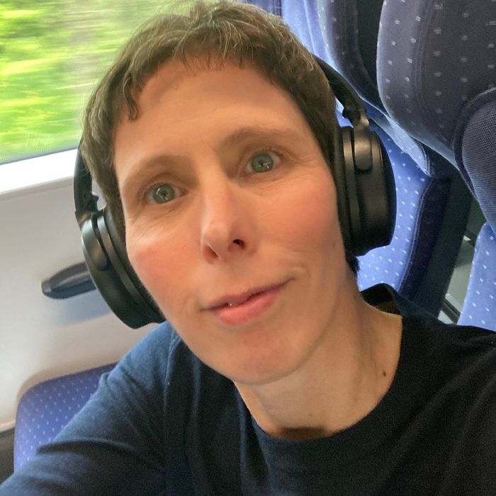 Portrait einer Frau mit braunen, kurzen Haaren. Sie trägt Kopfhörer und sitzt in einem Zug.