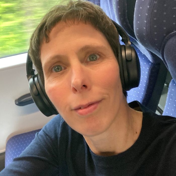 Portrait einer Frau mit braunen, kurzen Haaren. Sie trägt Kopfhörer und sitzt in einem Zug.