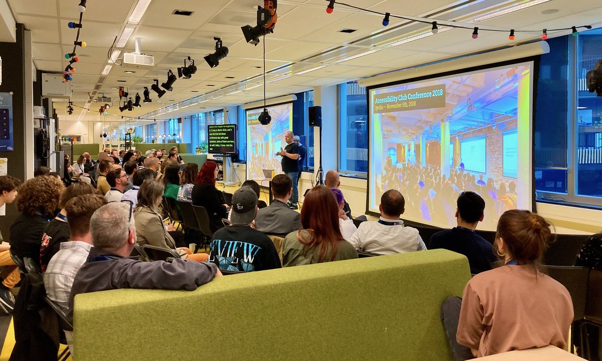 Blick über ein Publikum hinweg auf zwei Leinwände und einen Monitor mit Live-Untertitel, ein Mann spricht zum Publikum