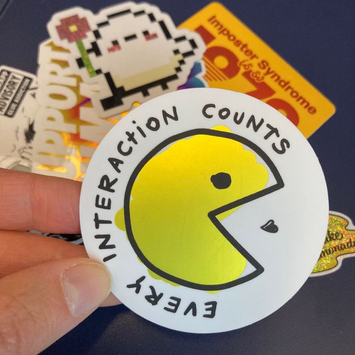 Verschiedene Sticker im Hintergrund. Eine Hand hält einen Sticker mit einer gezeichneten Pack Man Figur, drum herum steht geschrieben: Every interaction counts