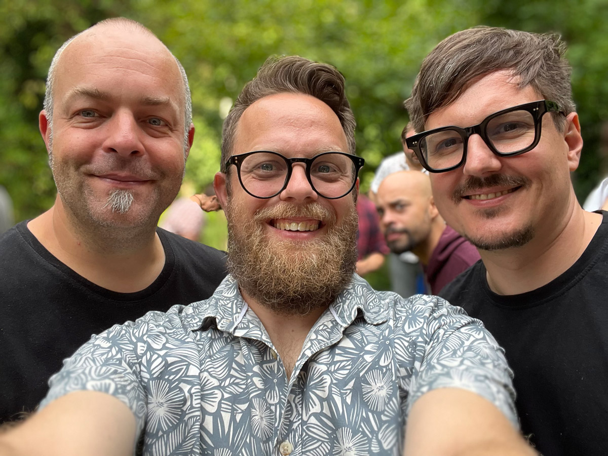 Oberkörper-Selbstportrait von Joschi, Andi und Markus. Alle lachen in die Kamera, die von Andi mit ausgestreckten Händen gehalten wird. Alle haben Bärte und tragen sommerliche T-Shirts bzw. Hemden. Andi und Markus tragen schwarze Brillen.