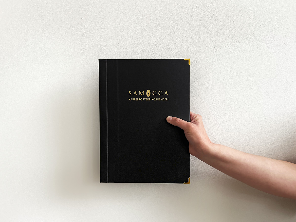 Die Speisekarte mit dem Titel &quot;SAMOCCA - Kaffeerösterei, Café, Deli&quot; wird von einer Hand vor eine weiße Wand gehalten. Der Einband ist schwarz, die Schrift in Gold geprägt.