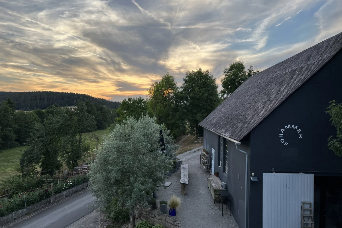 Abendstimmung am Hammerhof: Eine schwarze Scheune mit weißem "Hammerhof"-Schriftzug an der Fassade, umgeben von Bäumen und Wiesen, unter einem Himmel mit dramatischen Wolken und Sonnenuntergang.