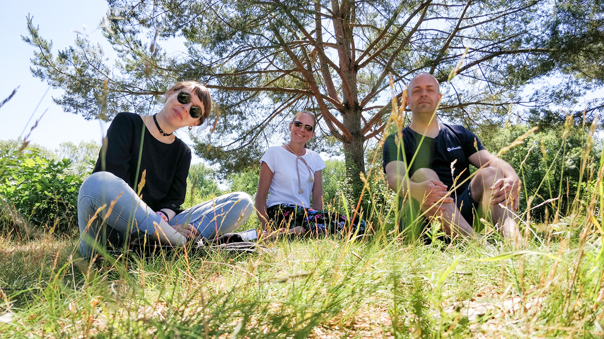 Nina, Annika und Joschi sitzen im Gras. Nina und Annika tragen Sonnenbrillen, im Hintergrund eine große Kiefer.