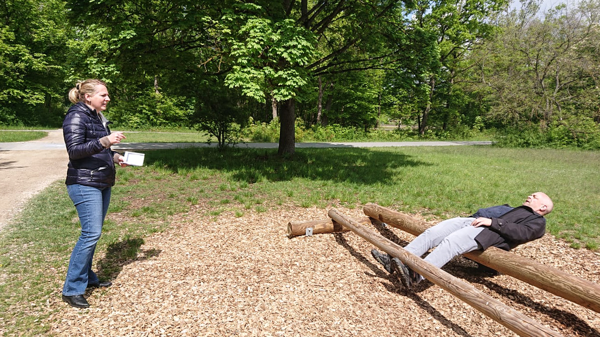 Zsuzsa stehend im Park vor einer Trimm-Dich-Anlage aus zwei horizontal montierten Baumstämmen. Joschi ist waagrecht zwischen den Stämmen eingehängt und macht Bauchmuskelübungen. Zsuzsa spricht gestikulierend mit ihm.