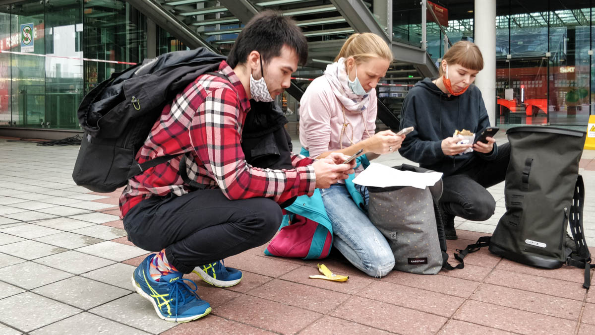 Jeff, Annika und Nina knien vor dem Flughafengebäude in München und nutzen ihre Handys