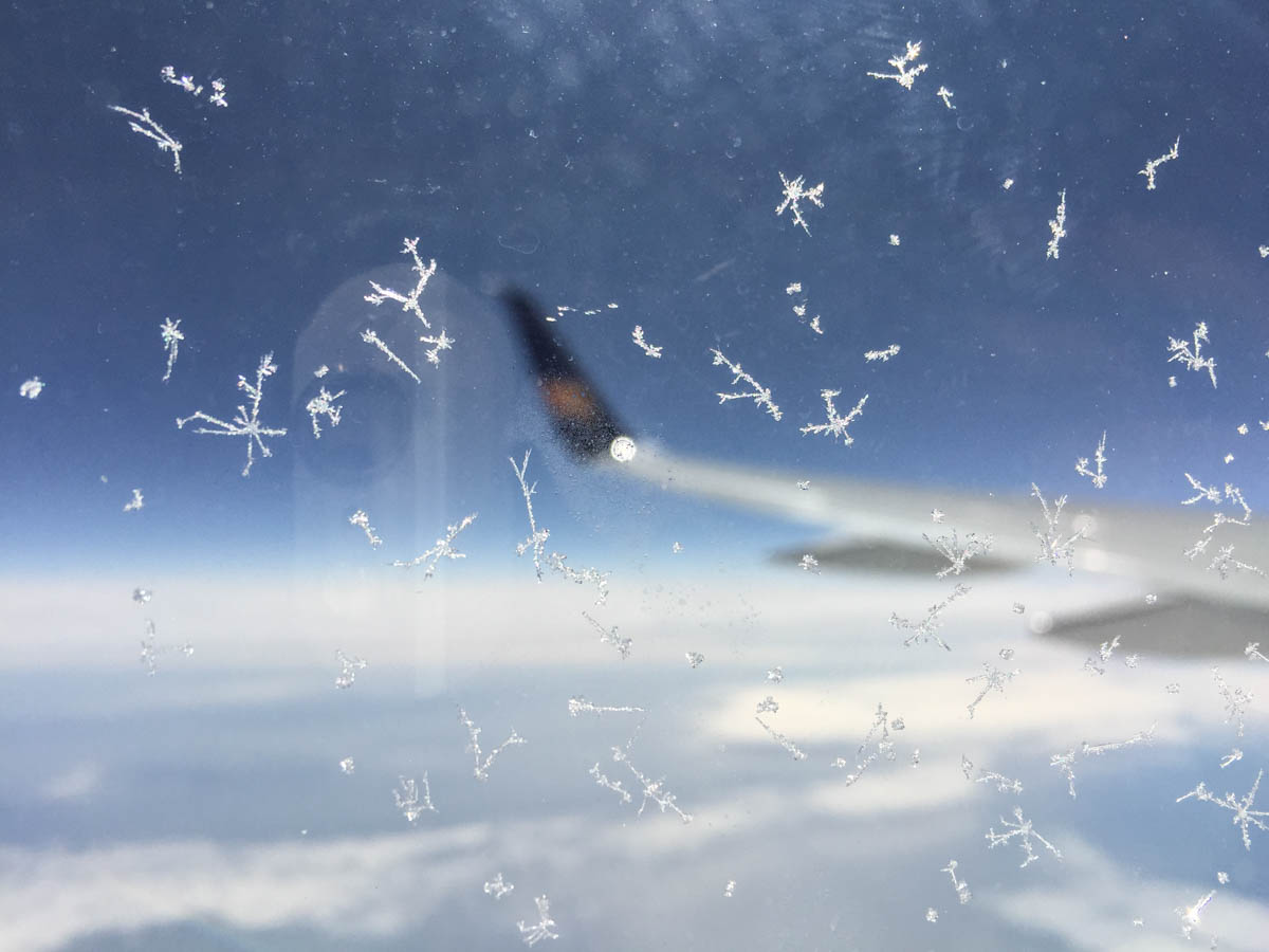 Eiskristalle auf einem Flugzeugfenster, im Hintergrund der Himmel und ein Flugzeugflügel