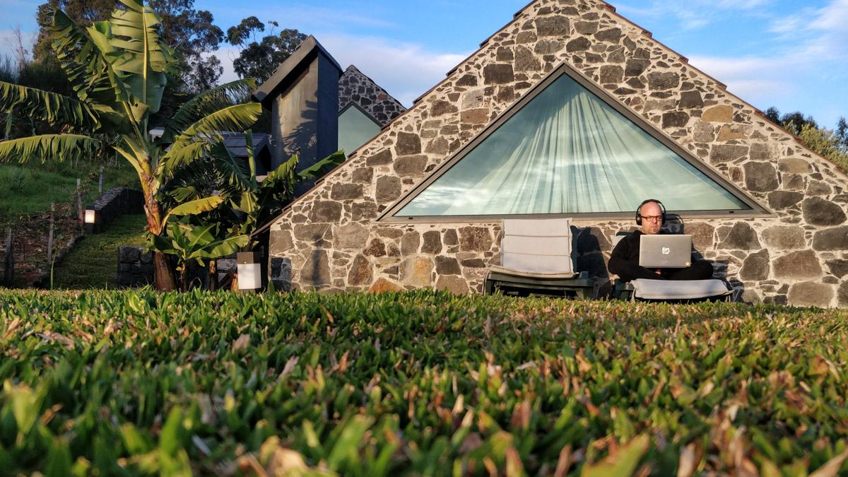 Joschi sitzt im Schneidersitz auf einer Gartenliege vor einem aus groben Steinen gemauerten Ferienhaus in Madeira. Er trägt einen Kopfhöhrer und arbeitet an seinem Laptop.