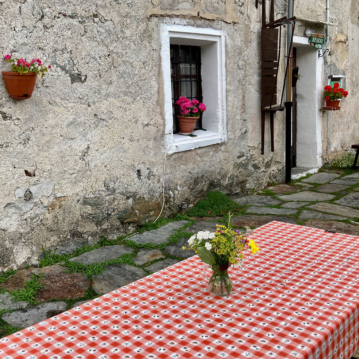 Ein Tisch mit einer rot-weiß-karrierten Tischdecke und einem kleinen Strauß Blumen steht vor einem einfachen, alten, alpin anmutenden Steinhaus.