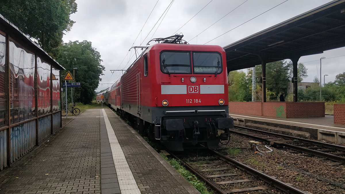 Der rote Zug, in dem Jakob sitzt, fährt in den Bahnhof Fürstenberg/Havel ein. Der Bahnsteig ist menschenleer. Der Himmel ist bewölkt.