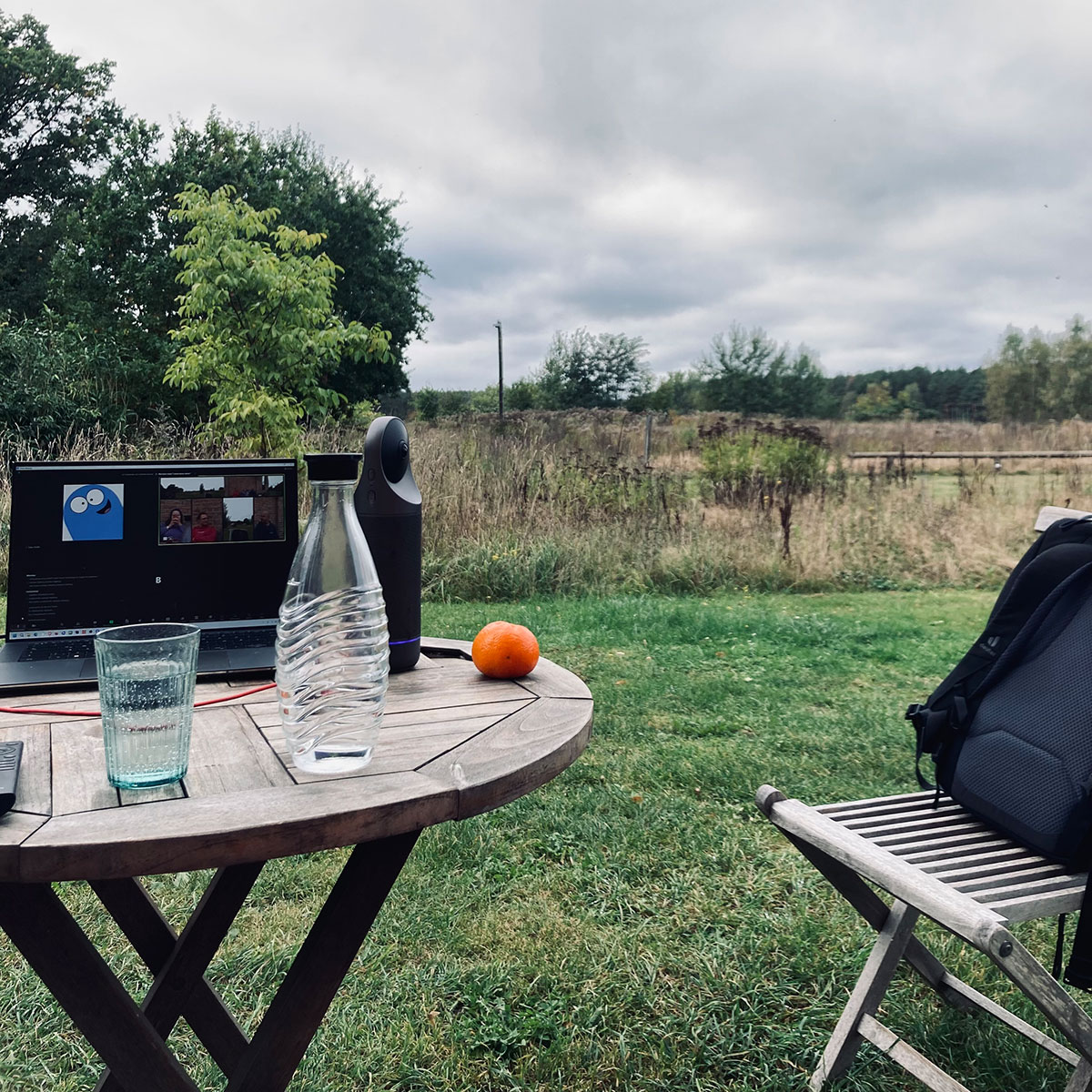 In einem großen Garten vor einem Feld steht ein runder Holztisch. Darauf ein Laptop mit einem aktiven Zoom-Meeting und eine Kamera. Davor Wasserglas, -flasche und Mandarine.