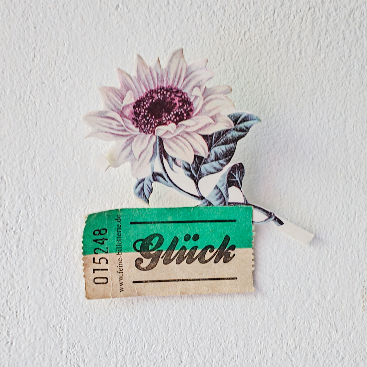 Kleine Collage an einer weißen Wand: Eine illustrierte Blume in zartem Rosa und eine weiß-grüne Papierwertmarke mit der Aufschrift „Glück".