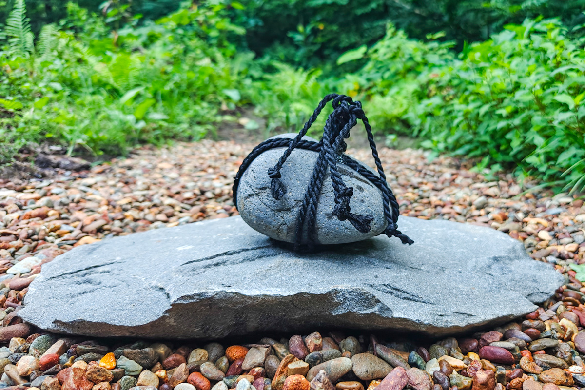 Ein runder grauer Stein, kunstvoll mit schwarzem Seil umwickelt, ruht auf einer flachen Schieferplatte inmitten bunter Kiesel. Im Hintergrund grünes Blattwerk.