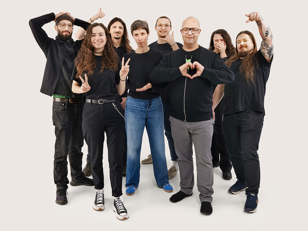 Das Tollwerk-Team vor hellbeigem Hintergrund, alle mit einer lockeren Haltung und unterschiedlichen Handgesten